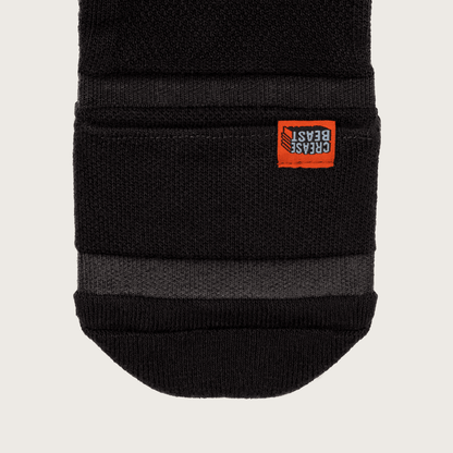 Close-up of a black Crease Beast Pocket Sock, showing the orange brand tag and a dark gray stripe.